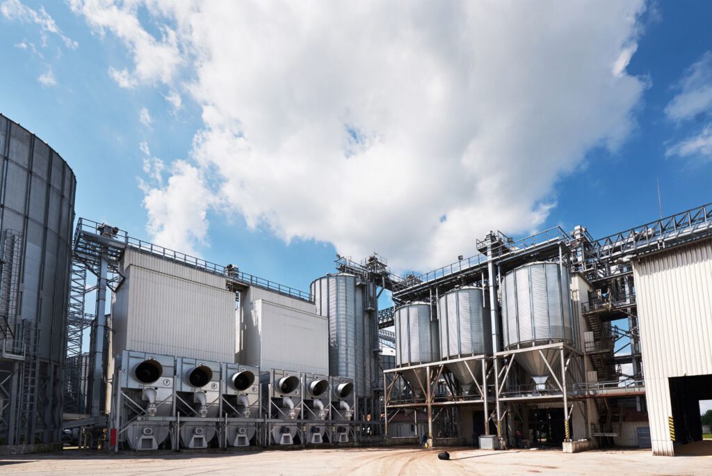 Planta industrial con grandes silos metálicos y maquinaria, cielo despejado con algunas nubes. Estructura moderna utilizada para almacenamiento y procesamiento de productos a granel. Ideal para ilustrar servicios de consultoría en inspecciones OCA y legalización EICI, asegurando el cumplimiento de normas y certificaciones en instalaciones industriales. Optimizado para atraer clientes interesados en la regulación y legalización de plantas industriales.