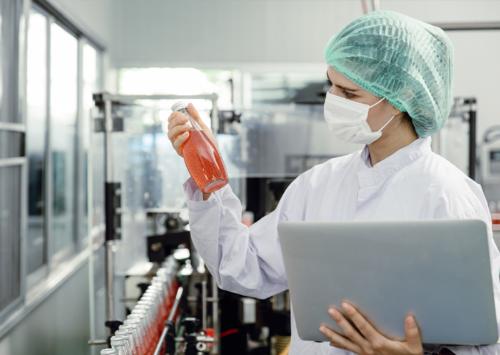 Consultoría en Seguridad Alimentaria, técnico inspeccionando botella en línea de producción con laptop y equipo de protección.